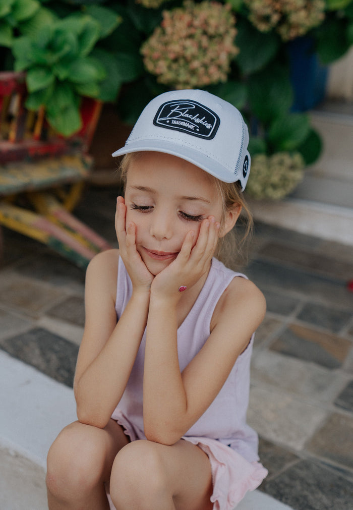 Young girl wearing a white cap with a logo, sitting on a stone floor.